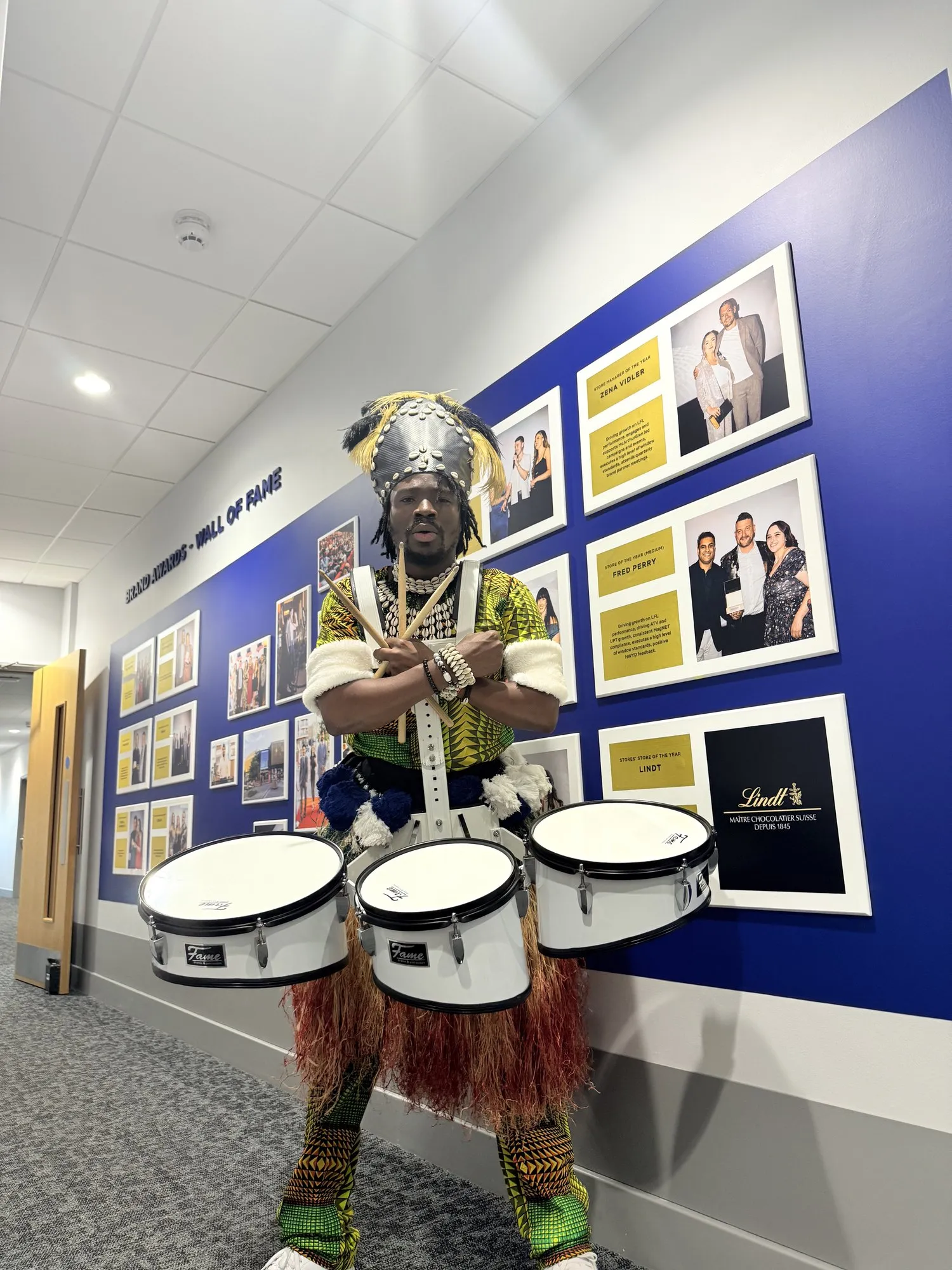 With three drums in a feathered costume beside a wall of fame